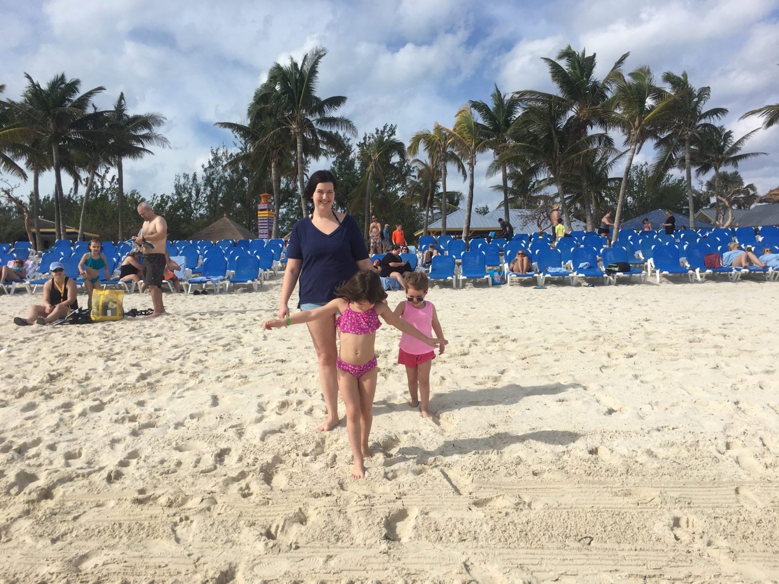 a mother and two young daughters walking on a tropical beach. Blue chairs are lined up behind them.
