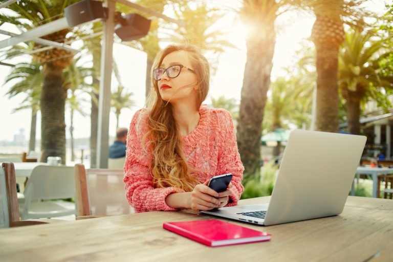 Attractive female freelancer hold smart phone while sitting at wooden table front open computer in modern coffee shop,young creative woman work on laptop while having breakfast on terrace, flare sun