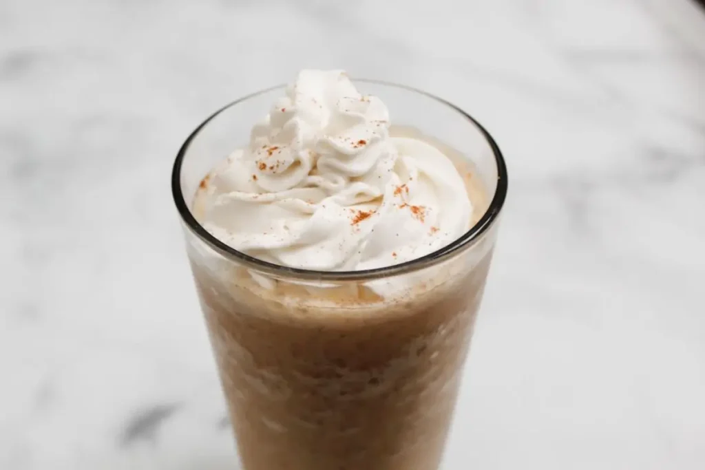pumpkin spice frappuccino with whipped cream on top in a glass cup on a white marble background