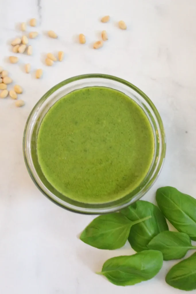 vegan basil pesto in glass dish surrounded by pine nuts and basil on marble surface