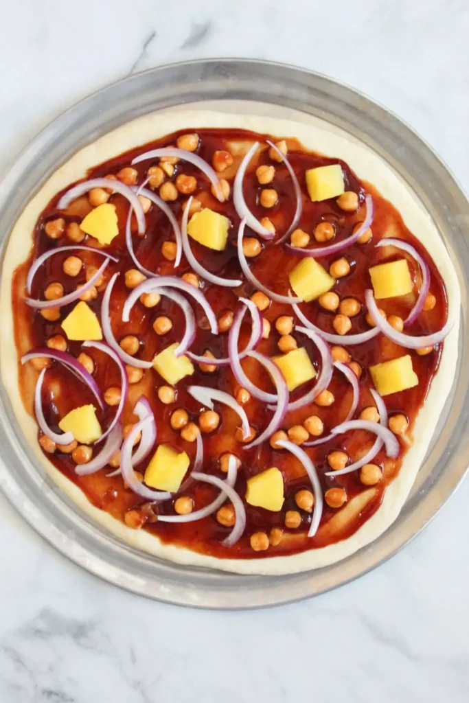 assembled pizza with bbq sauce, rolled out pizza, dough, chickpeas, pineapple, and red onions