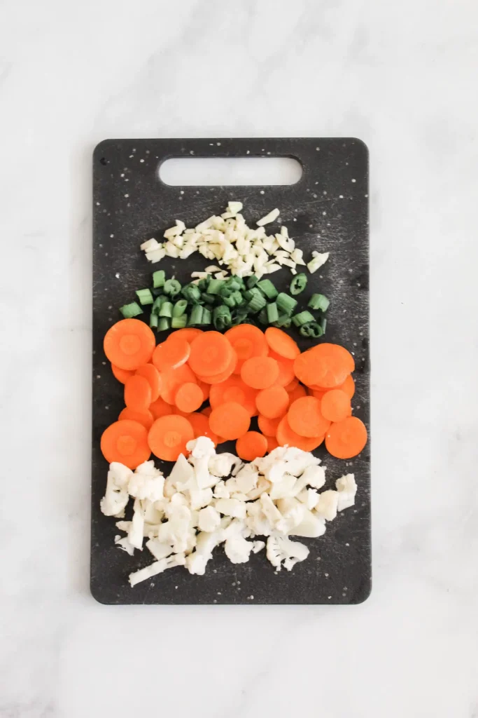 chopped up cauliflower, carrots, green onions, and ginger on black cutting board
